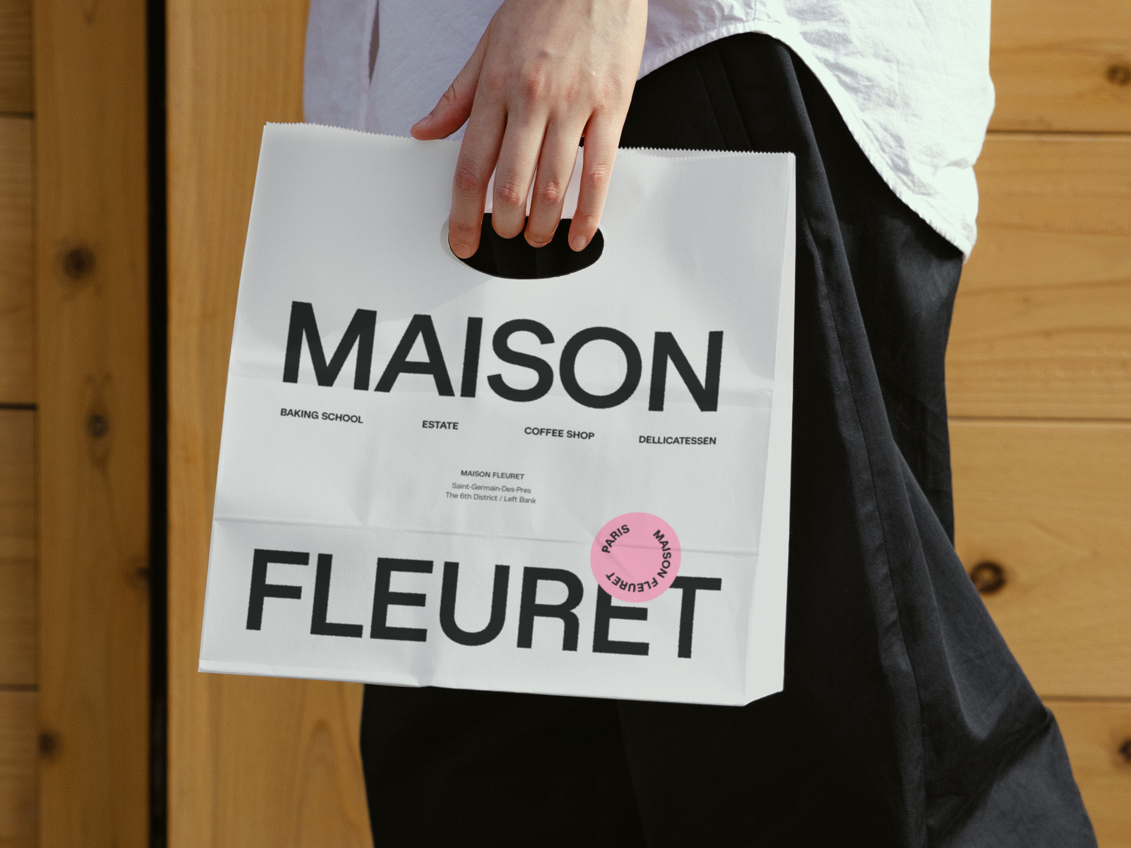 A white Maison Fleuret branded shopping bag held by a person, featuring black text labels, in front of a wood panel background.