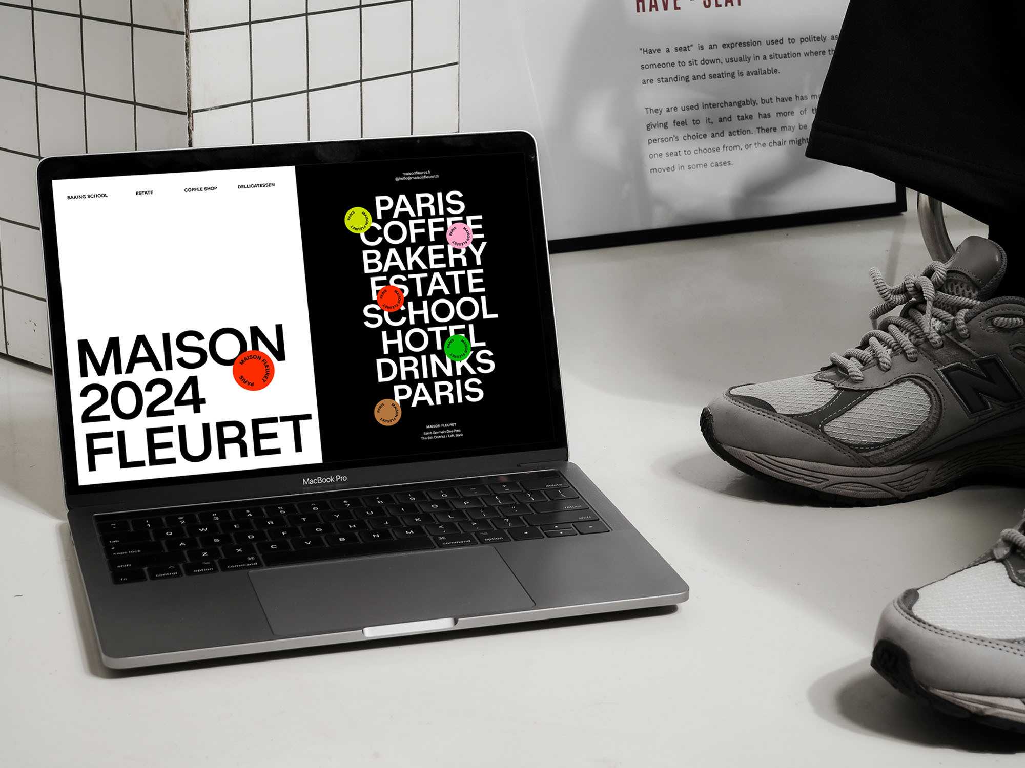 Laptop displaying website with 'Maison 2024 Fleuret' and list of locations, next to grey sneakers, against a white tiled wall and poster.