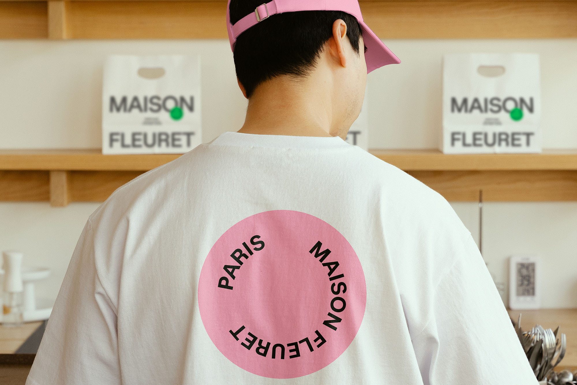 An adult with a pink cap wears a white Maison Fleuret t-shirt inside. Two shopping bags and kitchenware are visible in the background.