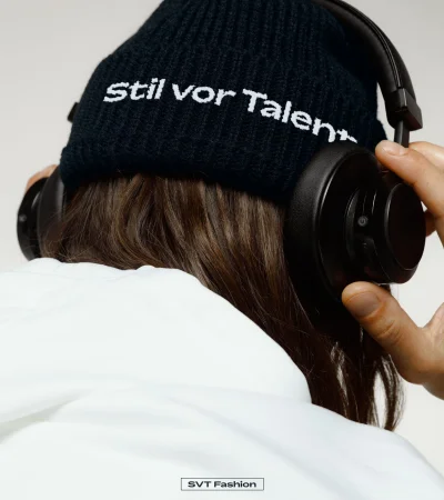 Person wearing a black knit beanie with embroidered white text and black over-ear headphones against a neutral background