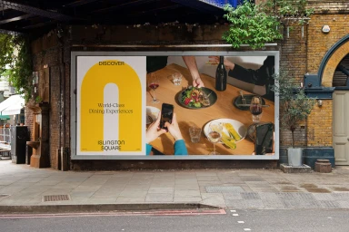 Outdoor billboard featuring yellow graphic branding and a dining photo mounted on an exposed brick building wall.