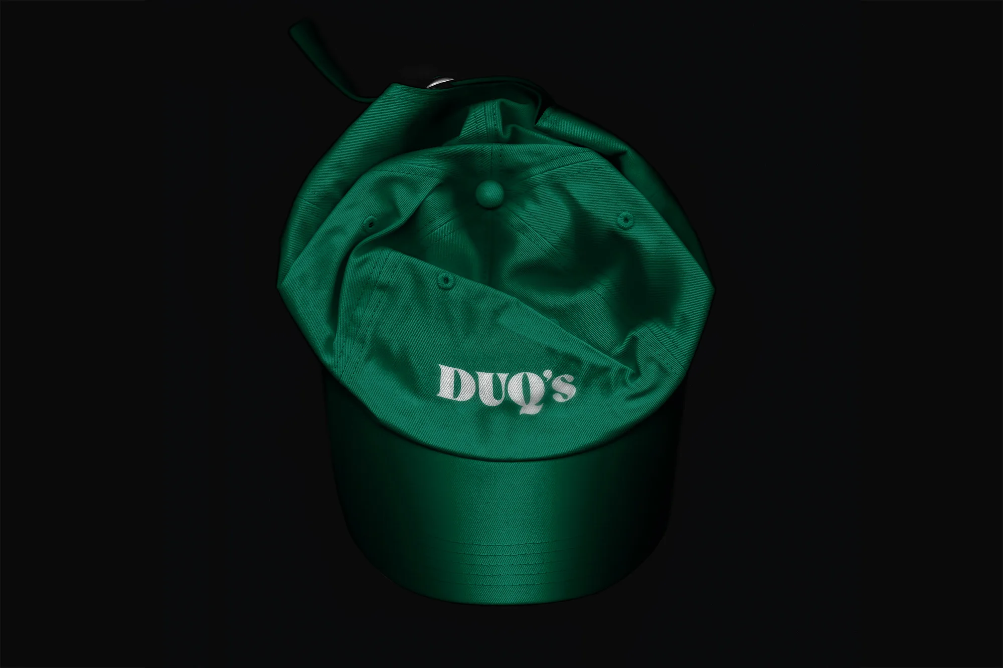 Overhead shot of a shiny, emerald green baseball cap with the name "DUQ'S" in white lettering, set against a stark black backdrop.