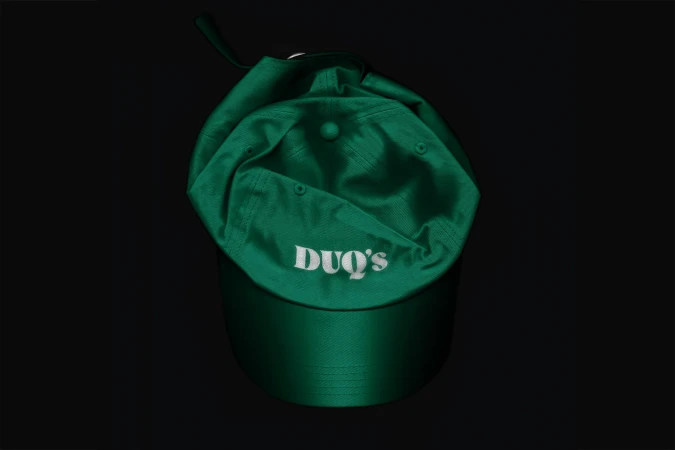 Overhead shot of a shiny, emerald green baseball cap with the name "DUQ'S" in white lettering, set against a stark black backdrop.