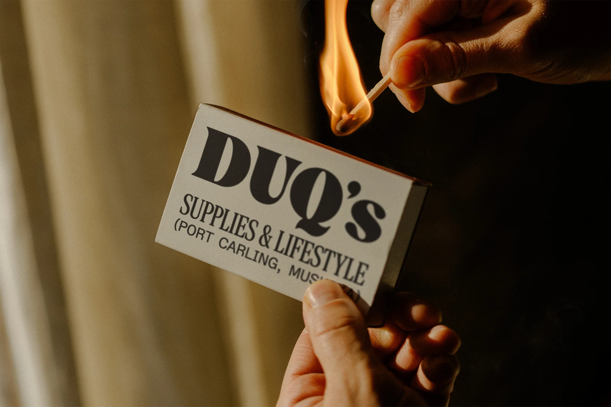 Close-up shot of a person lighting a match against the side of a matchbox with the words DUQ's Supplies & Lifestyle printed on it.