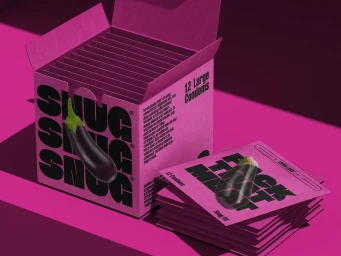An open pink SNUG condom box filled with foil packets and stacked packets beside it, featuring bold black typography and an eggplant graphic.