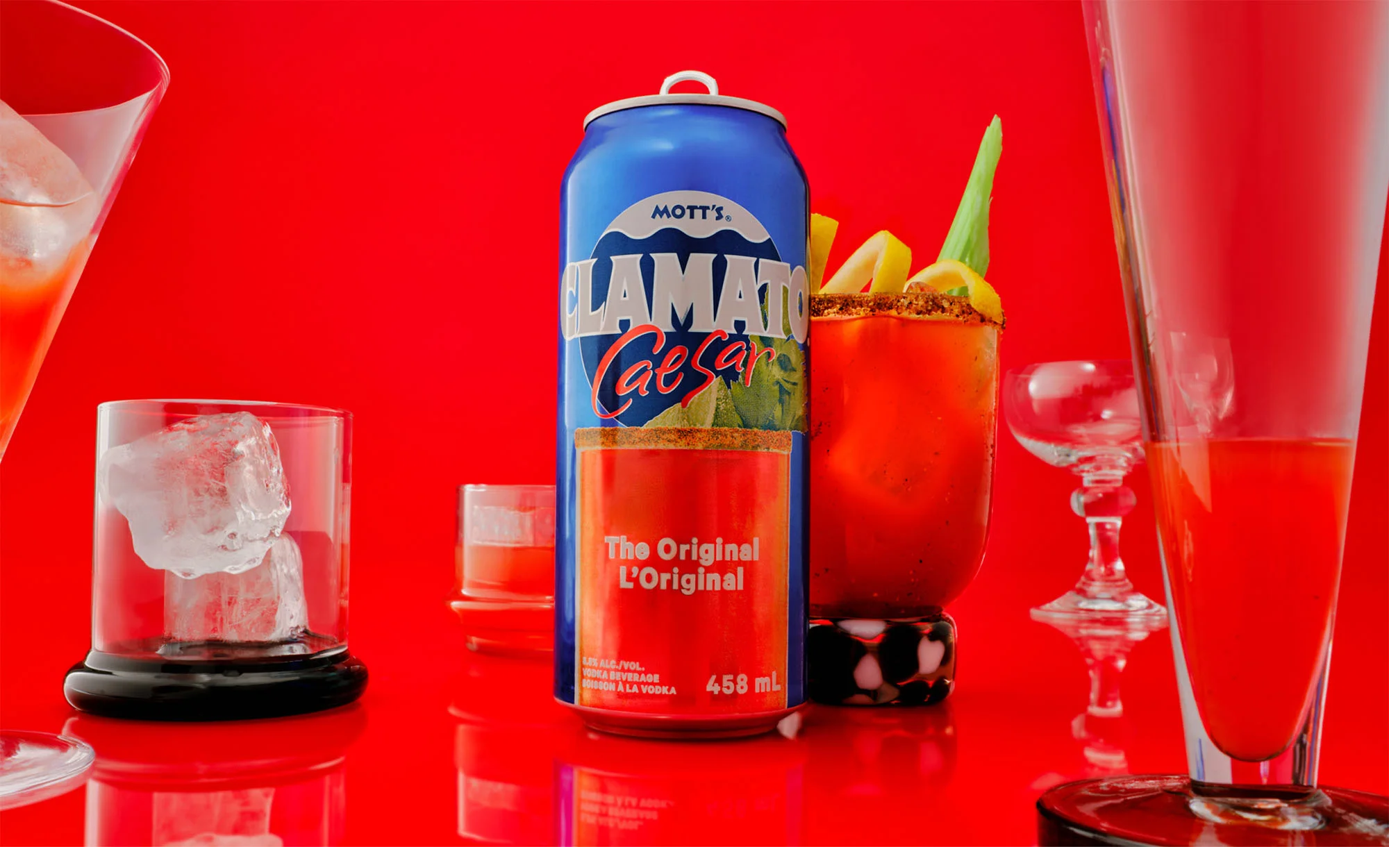 A vibrant studio shot displays a Mott's Clamato Caesar can surrounded by various glasses, some filled with a reddish drink, against a bright red backdrop.