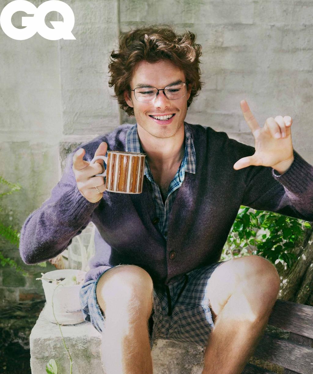 Smiling man in glasses, cardigan, and shorts sits on a bench, holds a mug, and gestures toward the viewer in an outdoor setting with a GQ logo.