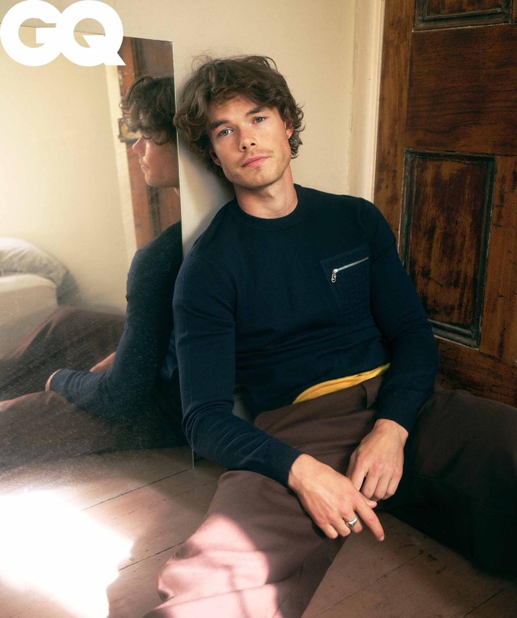 GQ portrait of a brunette man, indoors, leaning against a mirror next to a door; wearing a dark blue sweater and brown pants.