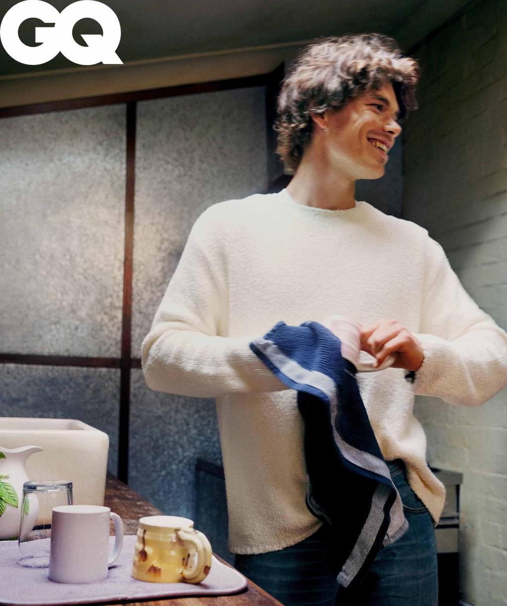 GQ magazine portrait of a smiling curly-haired man wearing a sweater and jeans, holding a towel in an indoor environment.