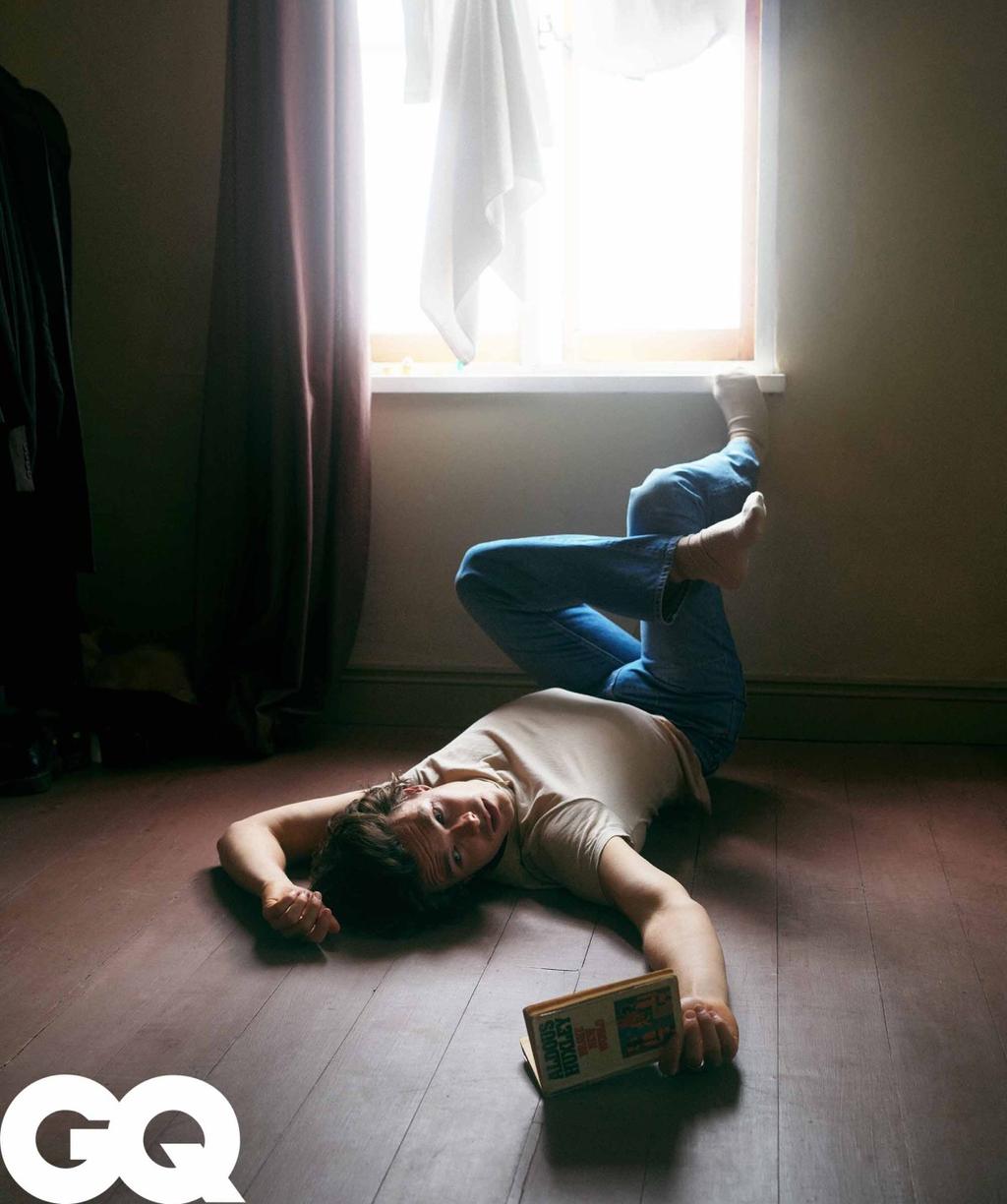 Adult male in casual clothing lying on a wood floor in a room with a book, posed near window light with a logo in the corner.