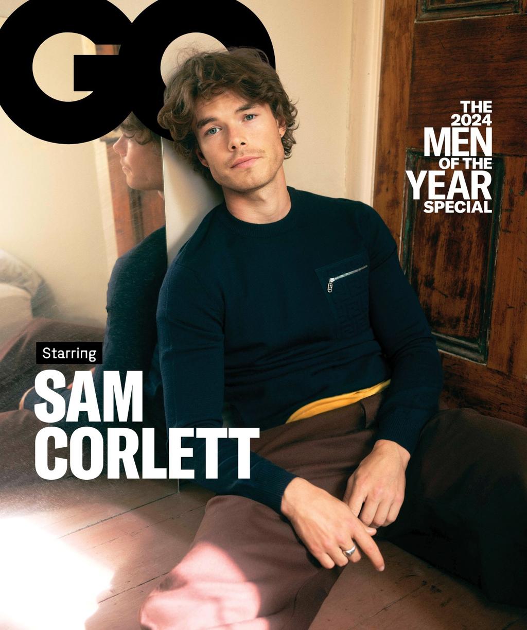 GQ cover featuring Sam Corlett in a navy sweater and brown pants, leaning against a mirrored wall with a wooden door in the background.