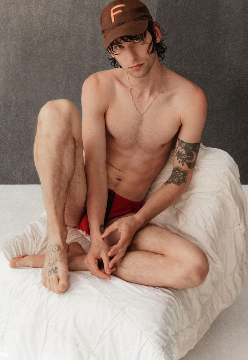 Shirtless adult male with baseball cap poses on bed, tattoos visible on arm and foot, against gray background in portrait orientation.