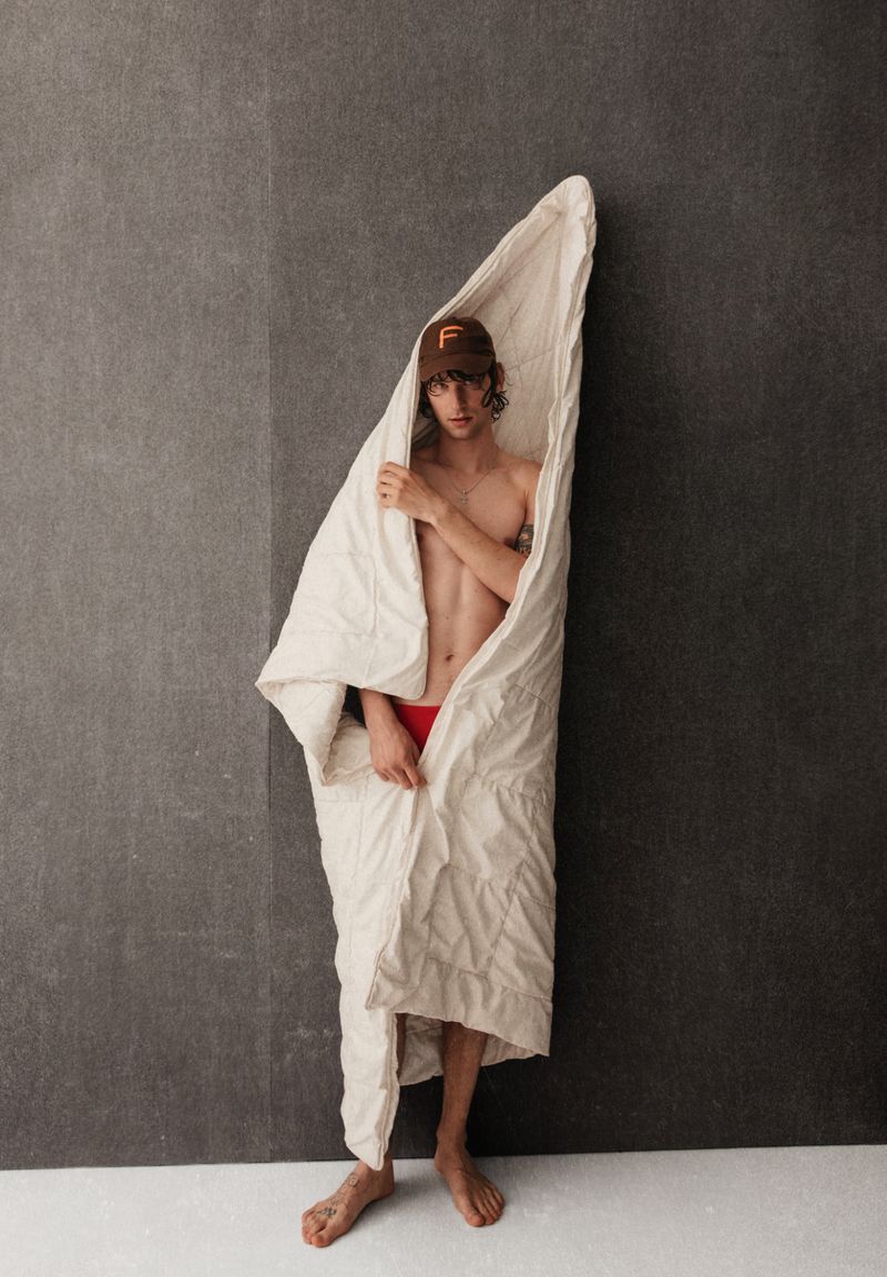 Young shirtless male model wearing a cap and wrapped in a white duvet standing against a gray wall in a studio setting.