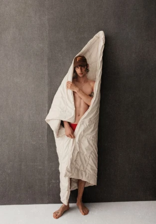 Young shirtless male model wearing a cap and wrapped in a white duvet standing against a gray wall in a studio setting.