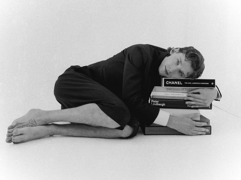 Adult male with short brown hair kneels, hugging a stack of books, in a full shot in a brightly lit indoor photo studio environment.