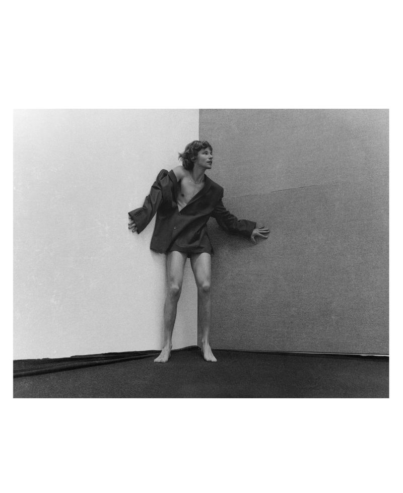 Black and white portrait of a slender person in an oversized blazer standing in the corner of a room with two-toned walls.
