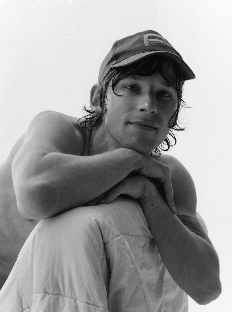 Black and white portrait of a young adult man with a cap, gazing at the camera, in an indoor setting with soft, natural light.