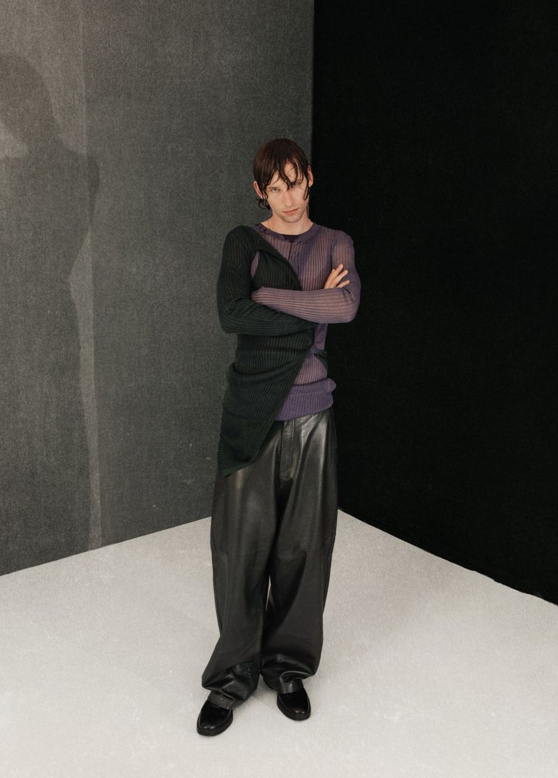 A slender adult male stands in a fashion pose, wearing a sheer purple long-sleeve shirt, dark green top, wide black pants, and black shoes.