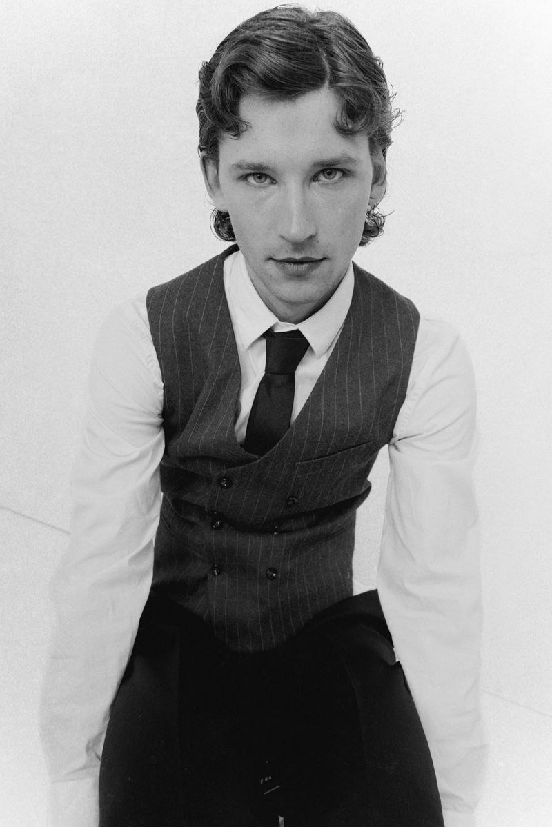Monochrome portrait of a young adult male wearing a pinstripe vest, white shirt, and dark tie, looking directly at the camera against a white backdrop.