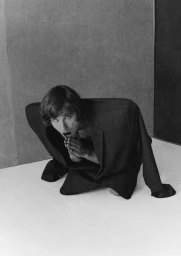 Model kneeling inside a structured oversized dark suit jacket with hands clasped in front, shot in black and white.