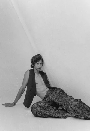 Monochrome fashion portrait of a male model seated against a plain white studio background wearing a vest and trousers