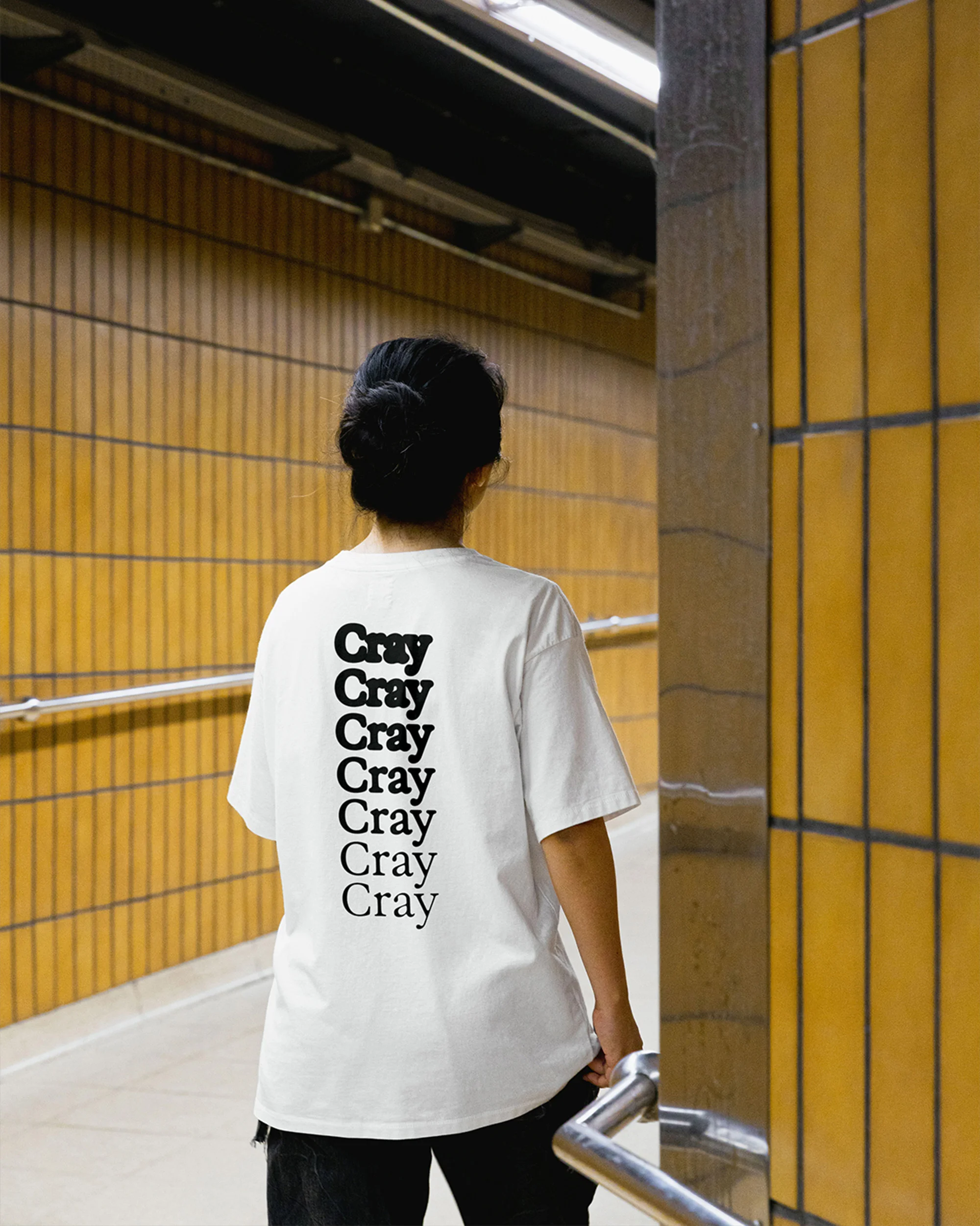 Back view of a woman in a white t-shirt with "Cray" printed in a repeating vertical design, standing in a tiled corridor.