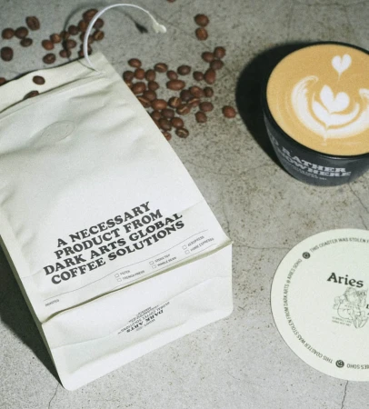 Overhead shot of a coffee bag, loose coffee beans, latte art in a mug, and Aries coaster on a concrete surface. 
