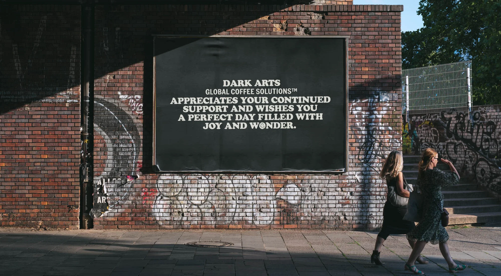 Two women stroll past a brick wall with a large advertisement for Dark Arts Global Coffee Solutions, adding a vibrant urban element.