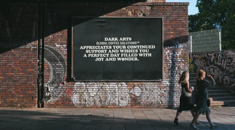 Two women stroll past a brick wall with a large advertisement for Dark Arts Global Coffee Solutions, adding a vibrant urban element.