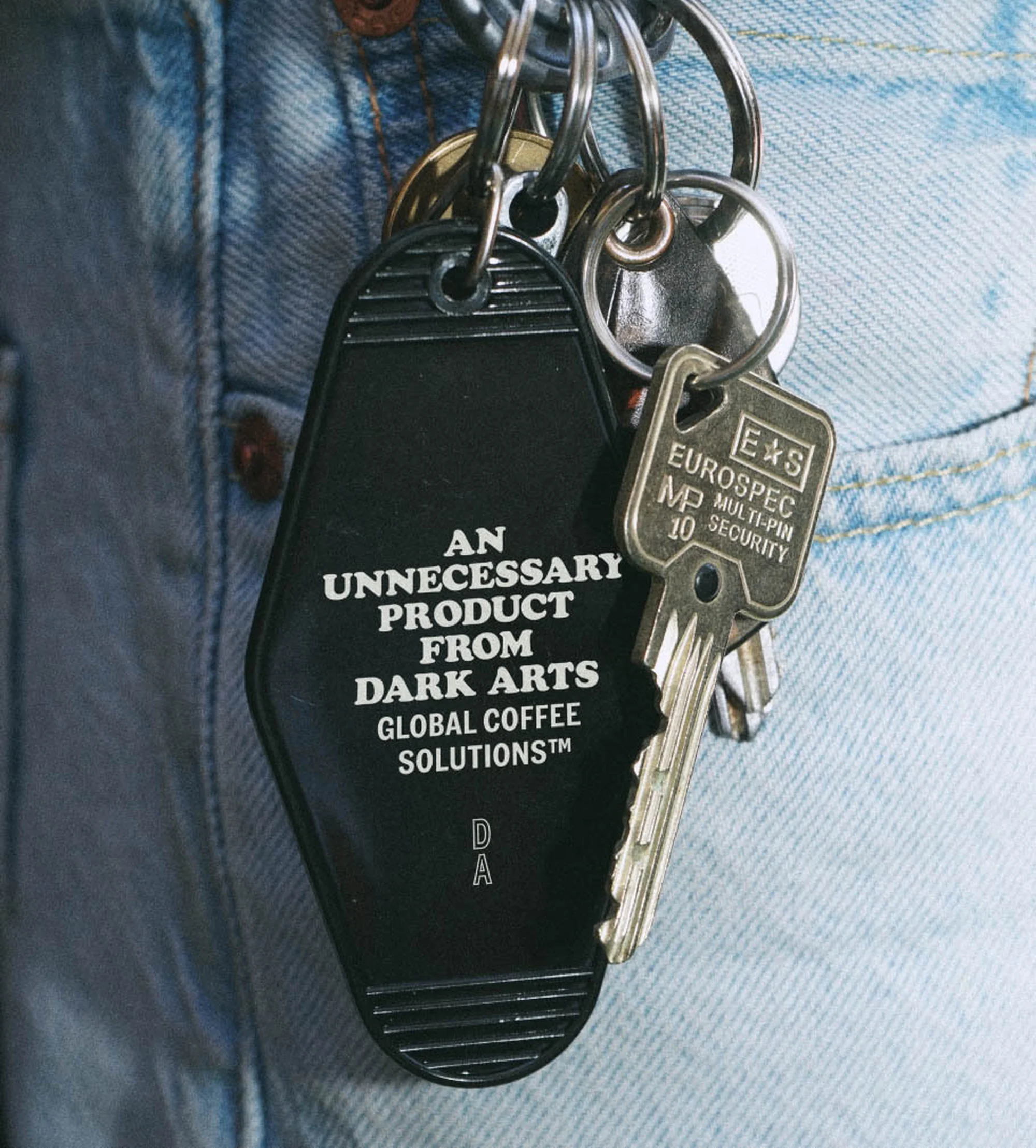 Close-up of jeans with a keychain featuring a black motel-style tag and Eurospec keys, highlighting dark arts-themed branding.