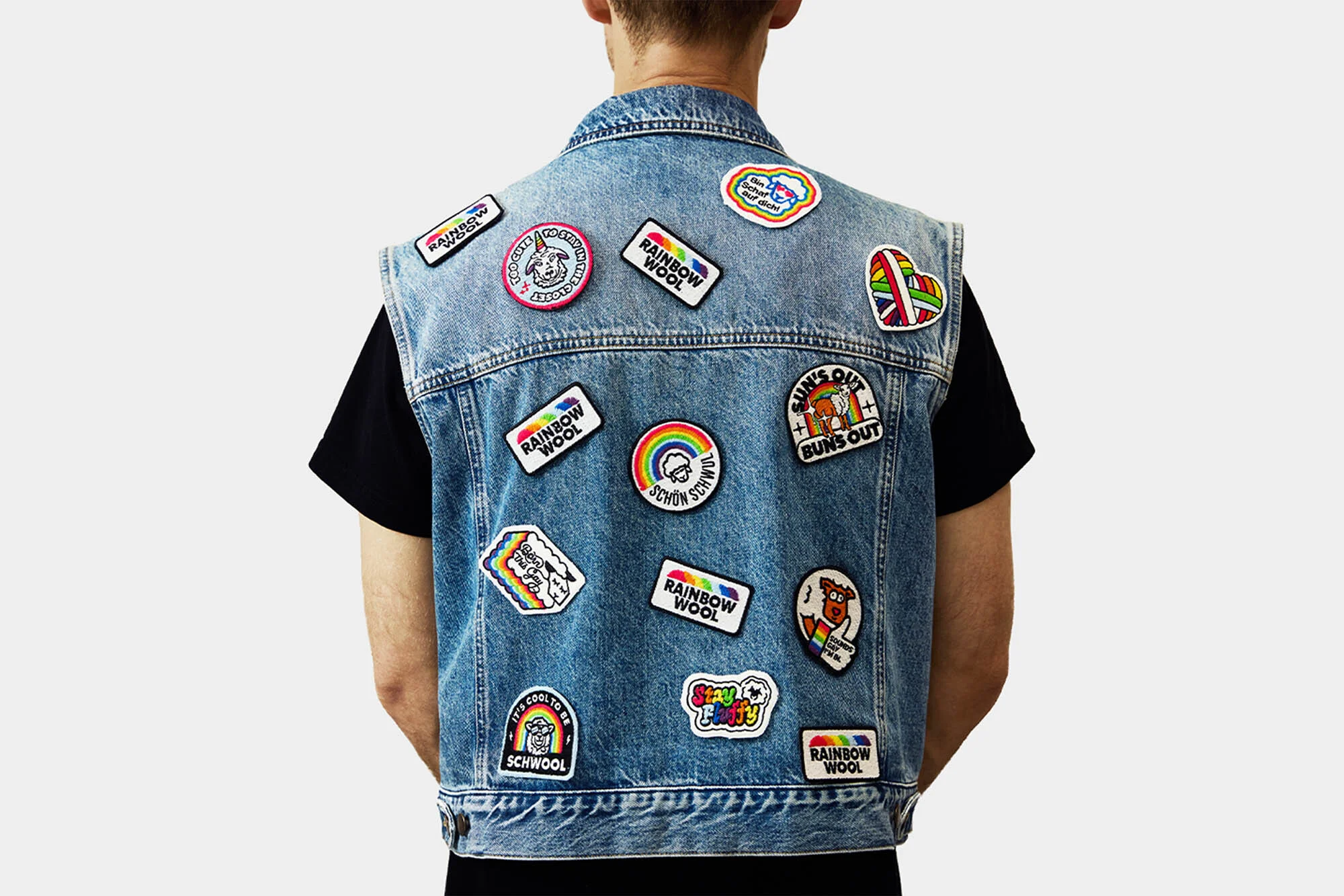 Back view of a person wearing a light blue denim vest covered in colorful patches against a white background in a studio.
