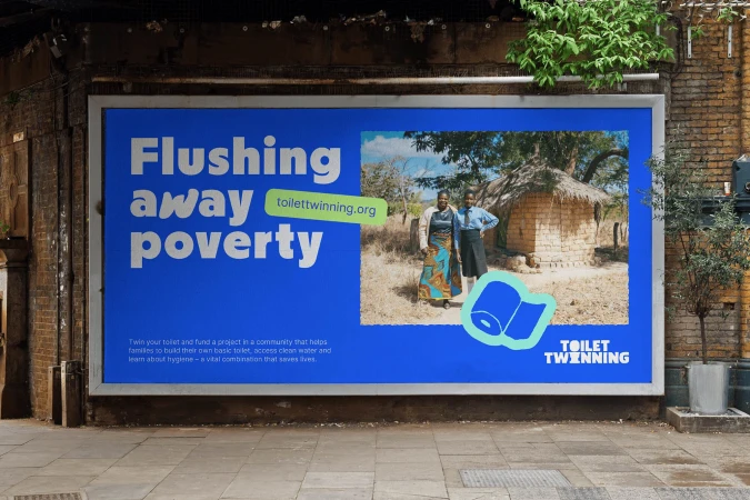 Outdoor billboard features two African women standing in front of a brick building with thatched roof. Text reads 'Flushing away poverty.'