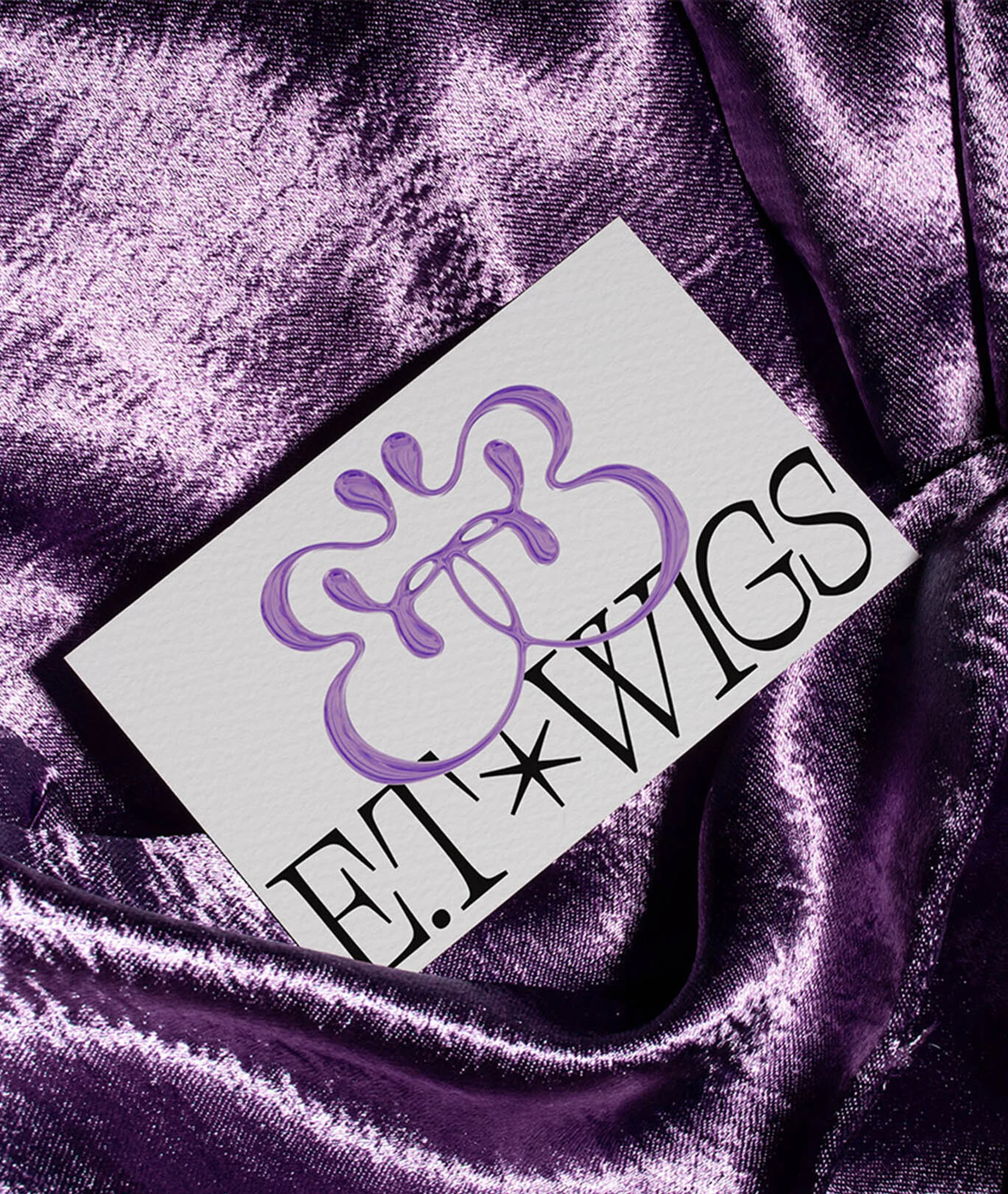Close-up of a business card with a purple logo and the text "E.J. Wigs" on a textured purple fabric background, product presentation.