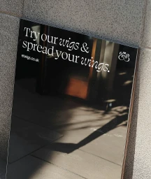 Black framed sign with white text reading Try our wigs and spread your wings resting against a textured concrete wall.