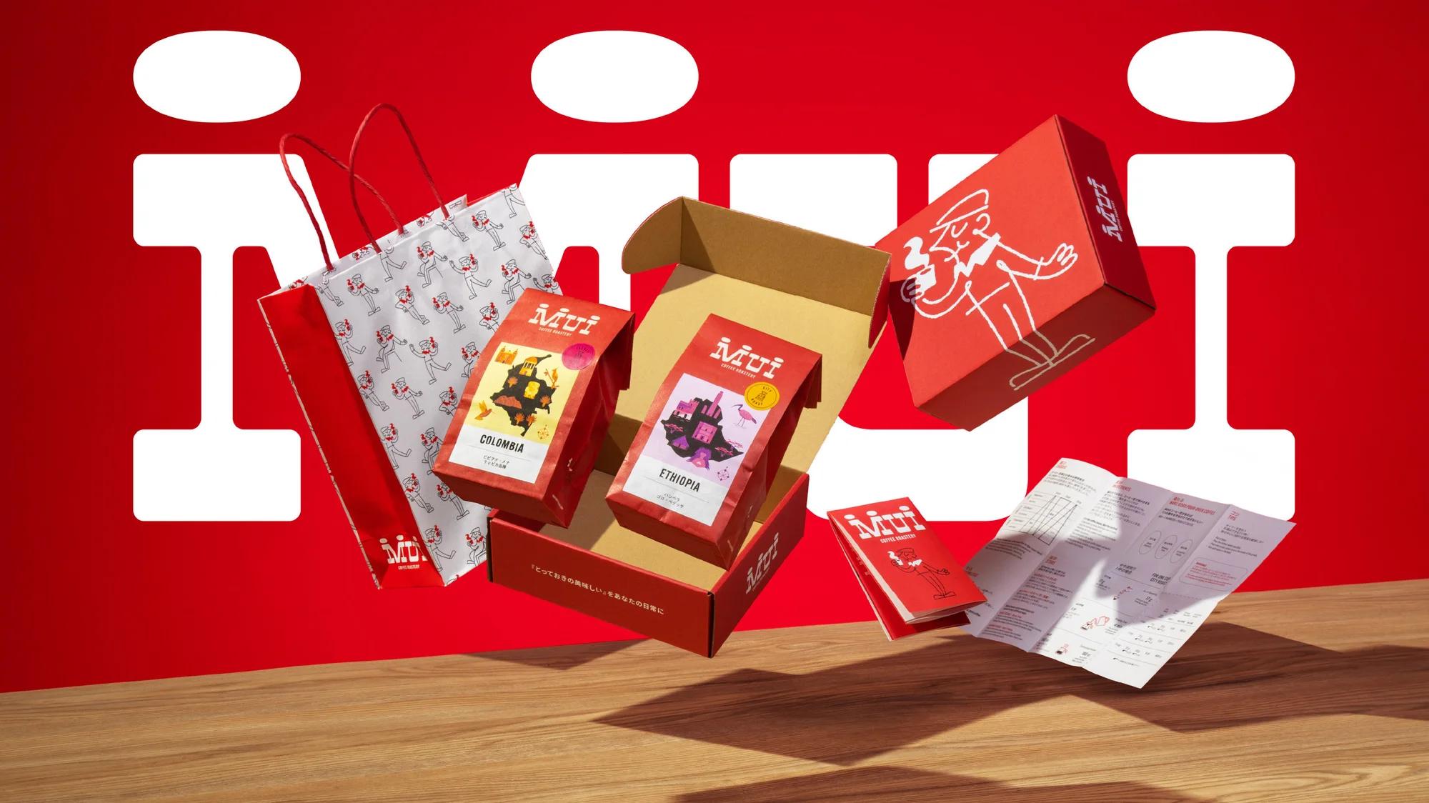 Still life shot of Mui coffee product packaging and promotional material including boxes, bags, and pamphlets against a red backdrop.