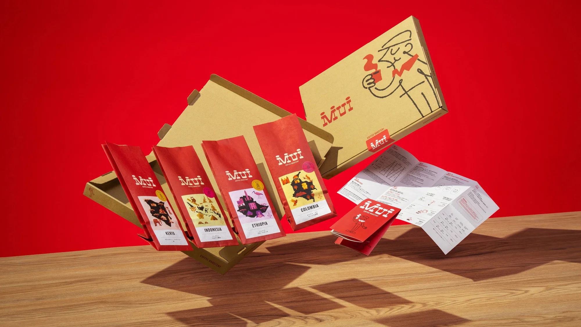 A gift box of Mui coffee bags is on a wood table against a red backdrop, featuring packages labeled with coffee bean origins.