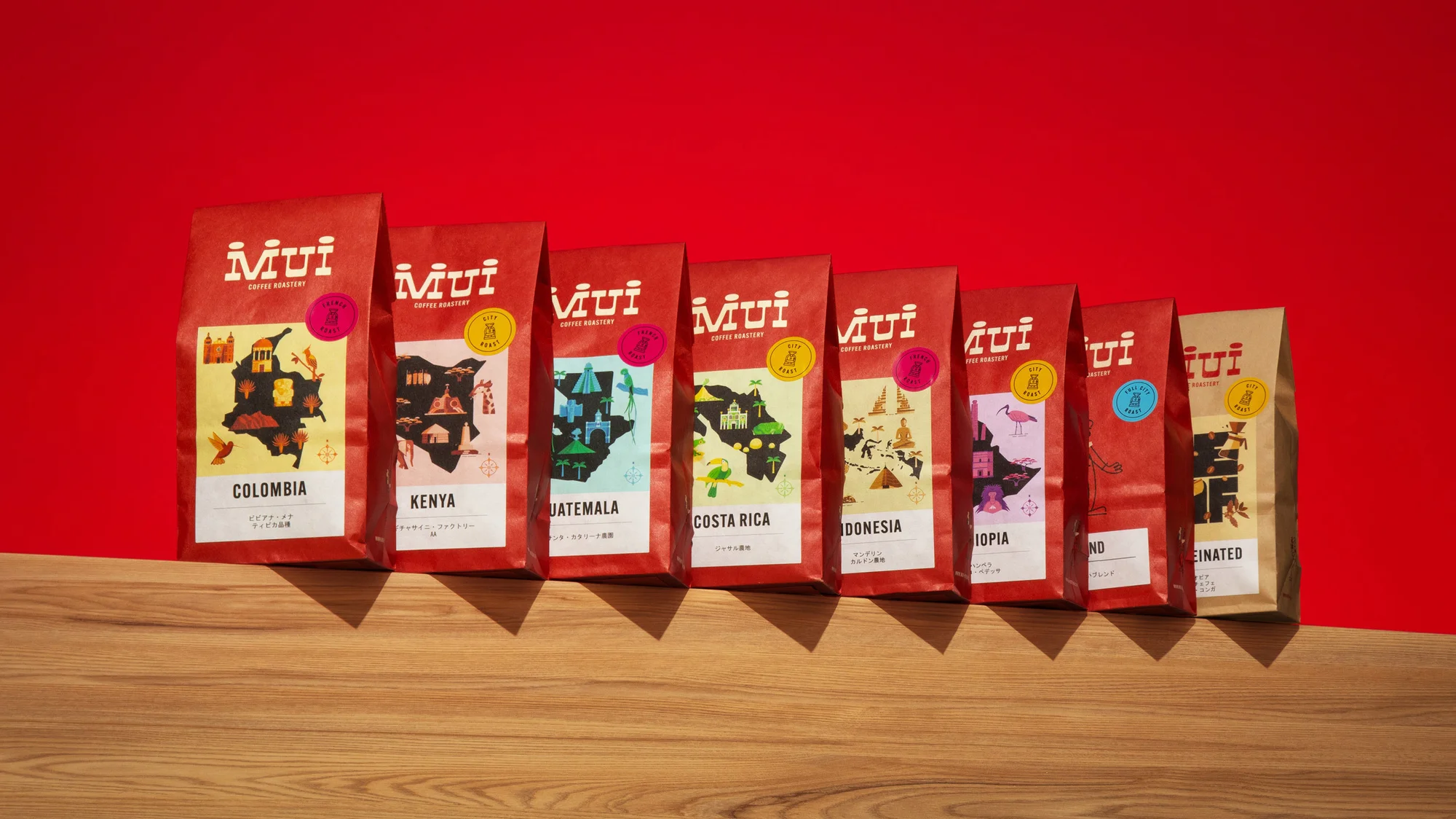 Eight colorful bags of Mui Coffee Roastery coffee beans, each labeled with a different country of origin, lined up on a wood surface.