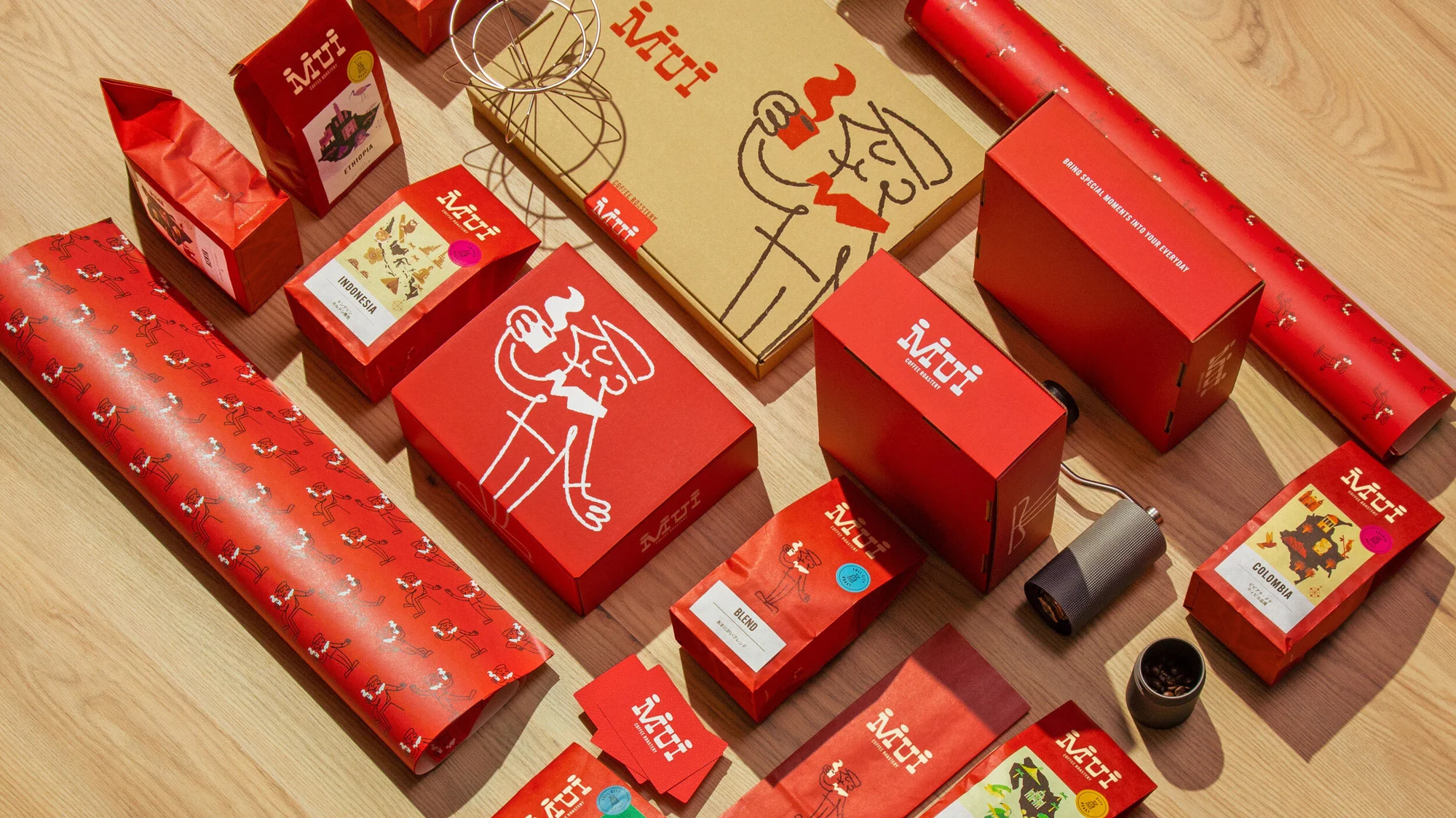 Overhead shot of Mui Coffee Factory boxes and packaging in red and tan, arranged on a light wood surface. Coffee bags and grinder.