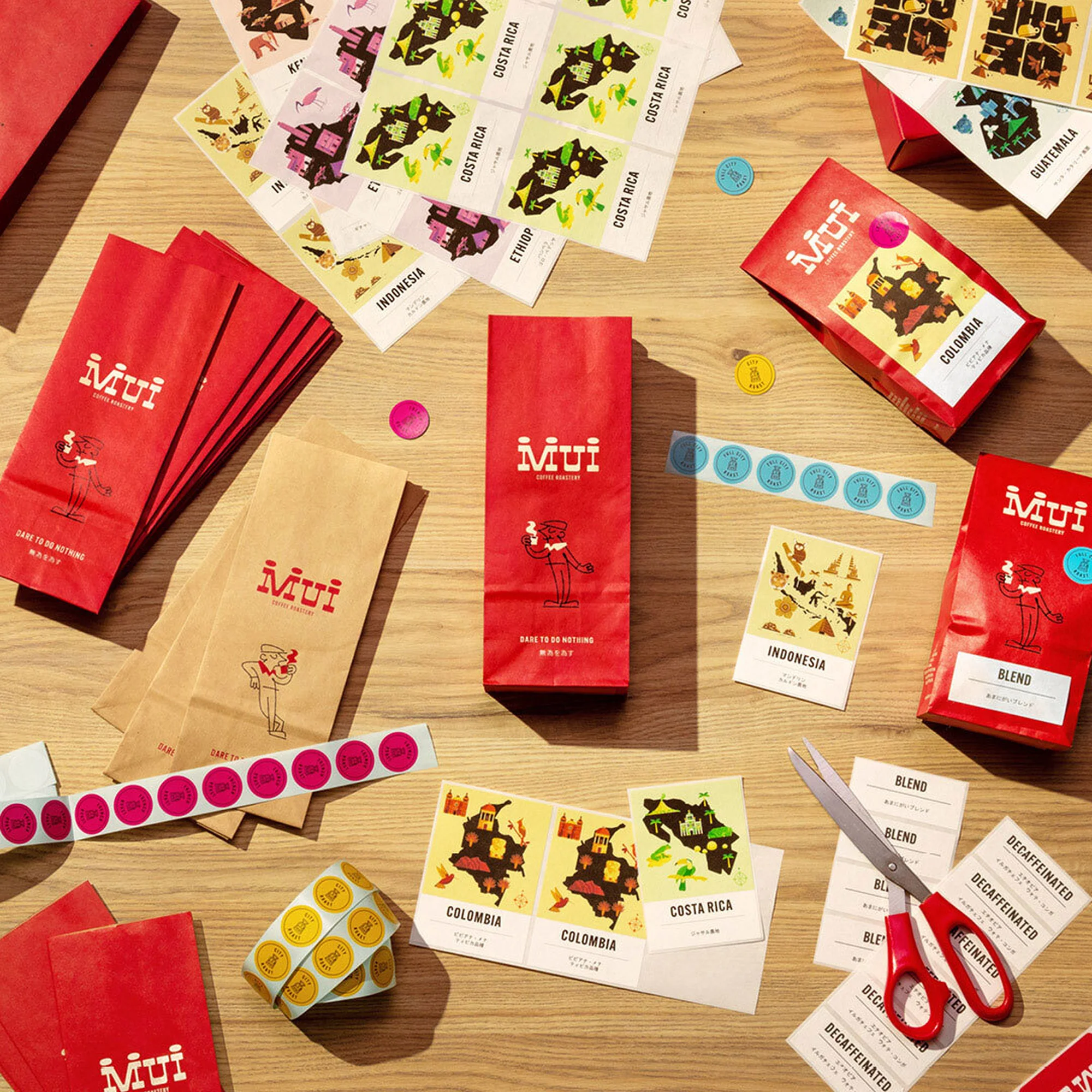 Overhead shot of Mui coffee packaging, including red and brown bags, stickers, and scissors, arranged on a light brown wooden surface.