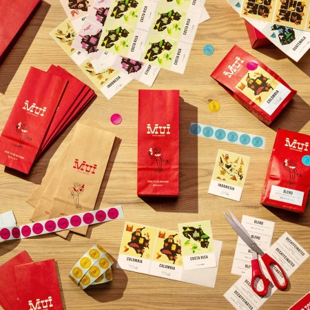 Overhead shot of Mui coffee packaging, including red and brown bags, stickers, and scissors, arranged on a light brown wooden surface.