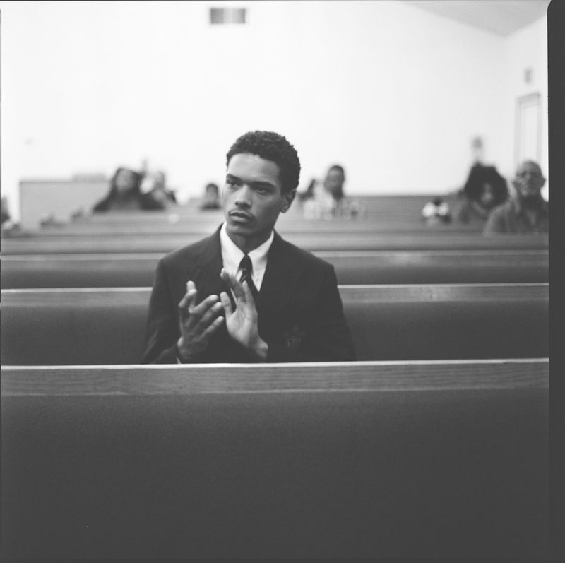 A monochromatic portrait of a young adult male in a suit clapping, sitting in a church pew with blurred people in the background.