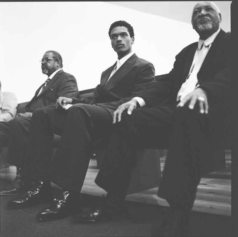 Three African-American men wearing suits sit indoors. The focus is on a young man in the center, with older men on either side of him.