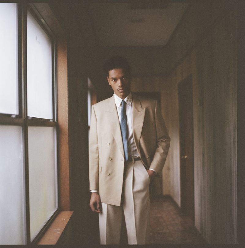 Young man in a tan double-breasted suit with a blue tie standing in a hallway next to a large window with natural light.