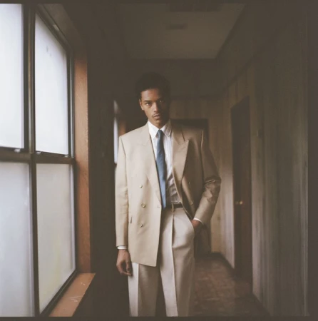 Young man in a tan double-breasted suit with a blue tie standing in a hallway next to a large window with natural light.
