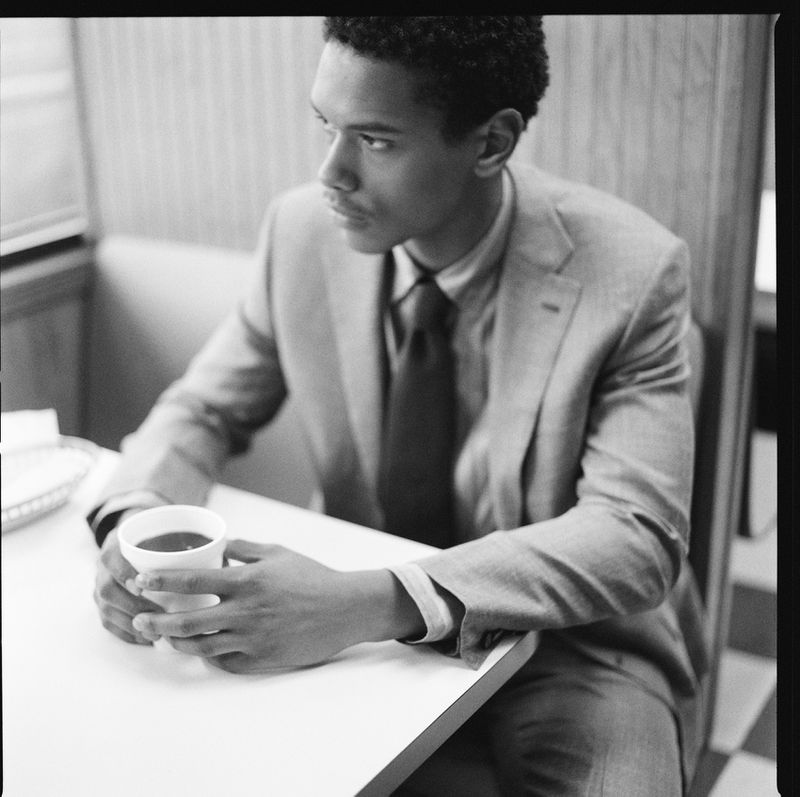 Black and white portrait of an adult man in a suit sitting at a table with a coffee cup, looking away in a softly lit interior.
