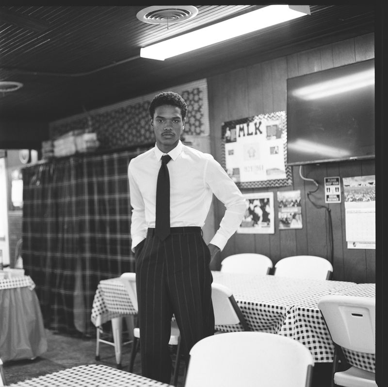 Black and white portrait of a stylish man in a white dress shirt and striped pants standing in a diner with gingham tablecloths.