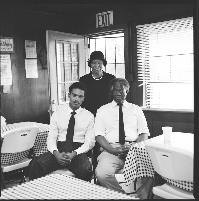 Monochrome indoor portrait of a seated group of three African-American adults, two men and one woman, dressed formally with checkered tablecloths.