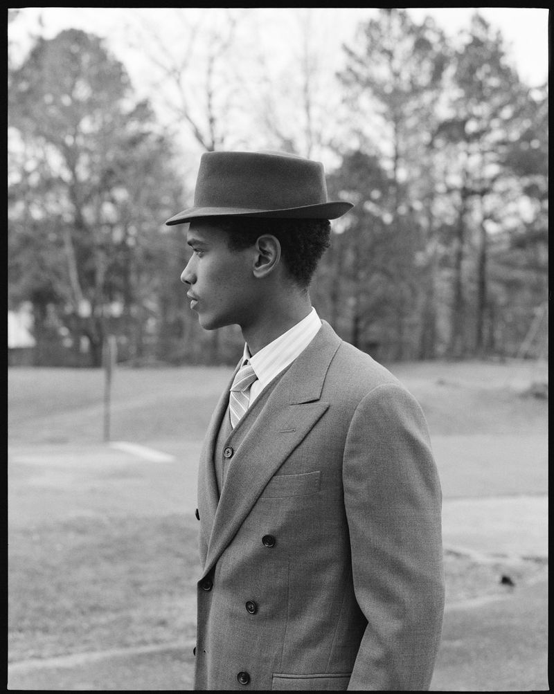 Monochrome portrait of a man in a suit and hat, looking to the side in an outdoor setting with trees and grass.