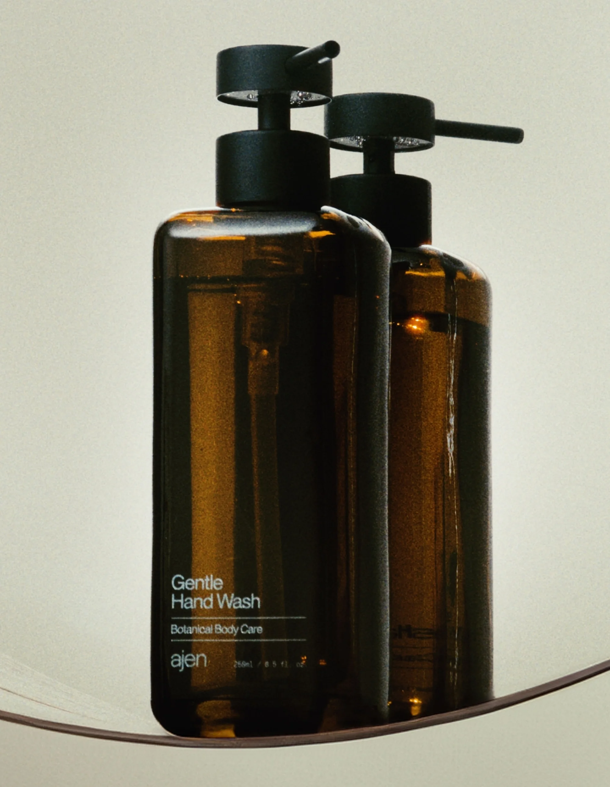 Two amber glass bottles with black pump dispensers sit on a curved surface against a neutral background. Minimalist design and body care focus.