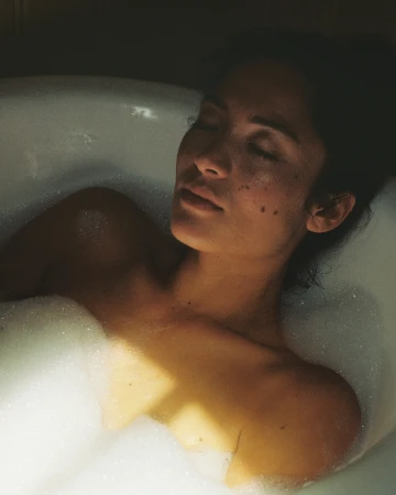 Adult female relaxing in a bubble bath, eyes closed, in a dimly lit bathroom setting with a vintage aesthetic and warm lighting.